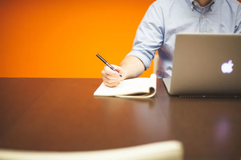 Person writing notes beside a laptop on a wooden desk