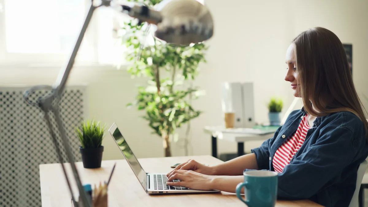 Woman smiling while working on a laptop at a home desk with plants and a coffee mug